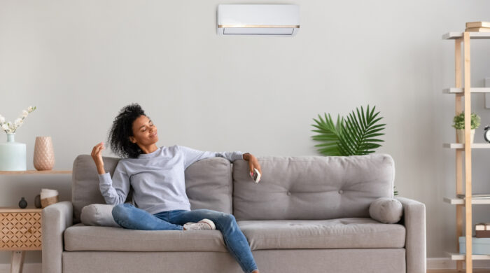African american relaxed woman sitting on comfortable couch in living room at modern home holds air conditioner remote control enjoying breathing fresh cool air at summer or warm air at winter season