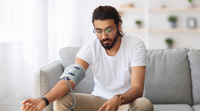 Long-haired, bearded millennial arab guy checking blood pressure at home, using tonometer while sitting on couch in living room, copy space. Handsome indian man suffering from hypertension