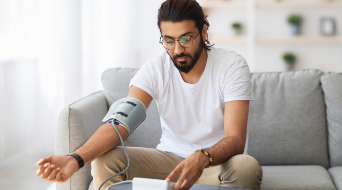 Long-haired, bearded millennial arab guy checking blood pressure at home, using tonometer while sitting on couch in living room, copy space. Handsome indian man suffering from hypertension