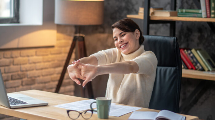 Smiling pleased female worker seated at the writing table stretching her fingers during the coffee break