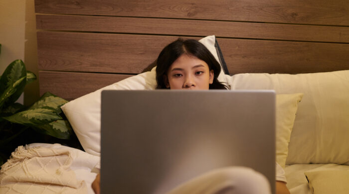 Tired young woman lying in bed with opened laptop, reading article or watching video