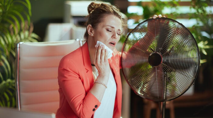Sustainable workplace. modern 40 years old bookkeeper woman in modern green office in red jacket with electric fan and napkin suffering from summer heat.