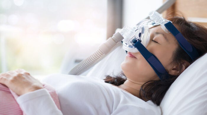 Senior woman using cpap machine to stop choking and snoring from obstructive sleep apnea with bokeh and morning light background.
