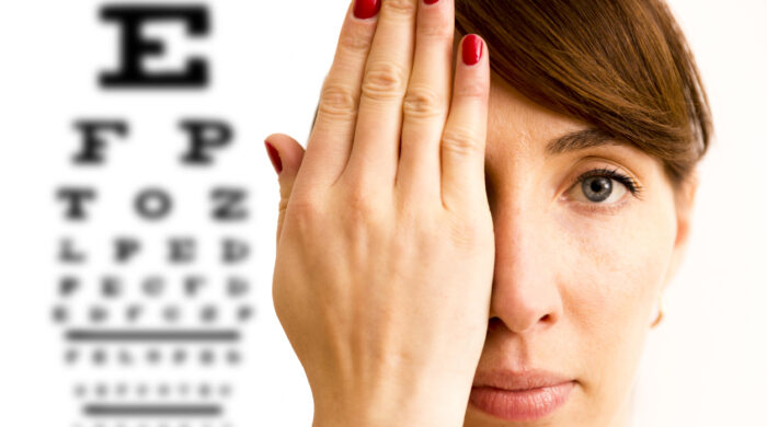 Young woman is covering his face with hand and checking his vision. Chart for eye sight testing in background.