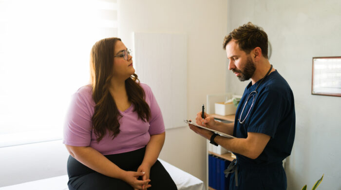 Plus size woman sitting on examination table while talking to doctor taking notes on clipboard during medical consultation in doctor's office